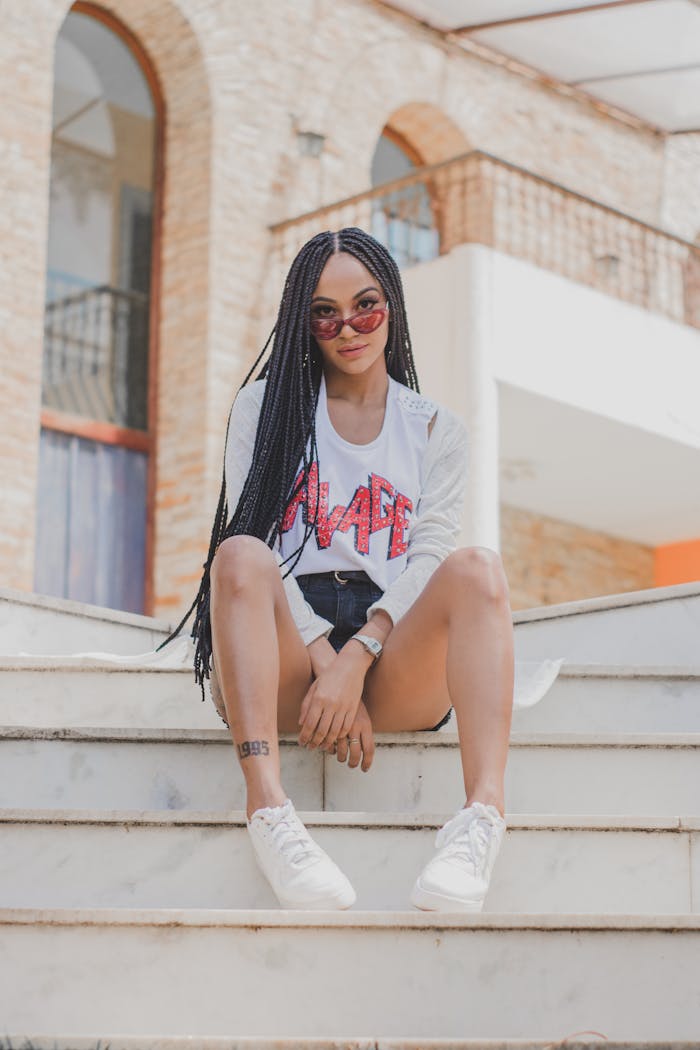 Fashionable young woman posing confidently on urban stairs with sunglasses and braids.